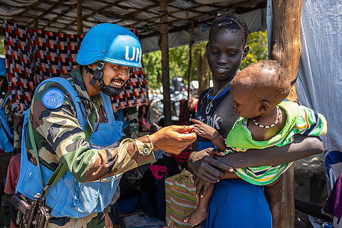 A man in blue UN body armour and helmet plays with a young child being held by a woman
